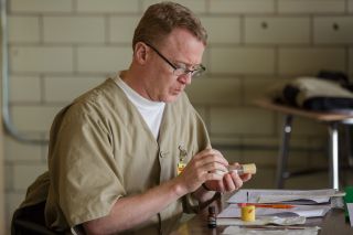 Eric Graves , a white middle aged man with sandy blonde hair , is sitting and working on a project for school with his hands. He is wearing a tan prisoner uniform.