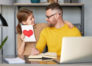 Daughter gives Father's Day card to father on Father's Day while he works on laptop computer,